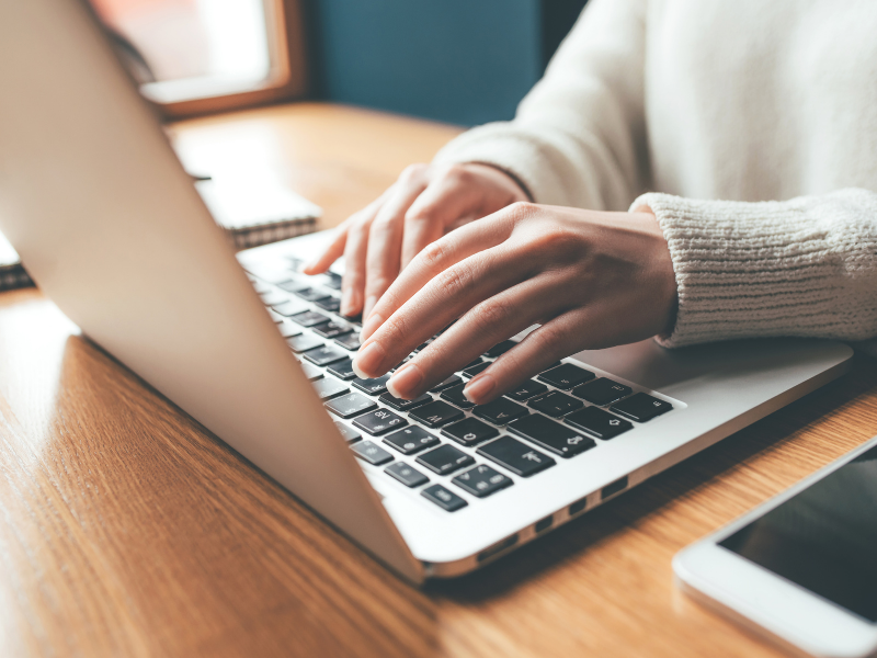 Händer som skriver på en dator. Bredvid datorn finns en mobil och ett anteckningsblock.